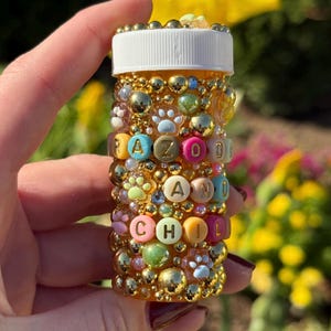 May include: A decorated pill bottle with the words "RAZOO AND CHILL" spelled out in colorful letter beads. The bottle is covered in gold beads, paw print charms, and other decorative elements. The bottle has a white cap.