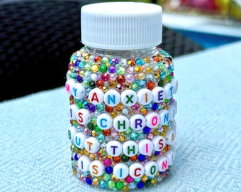 Puede incluir: Una pequeña botella de vidrio transparente con tapa blanca, cubierta de brillantes de colores. La botella tiene el texto "MY ANXIETY IS CHRONIC BUT THIS IS ICONIC" escrito con cuentas de letras blancas. Sobre una superficie azul claro.