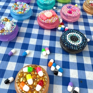 May include: A collection of bedazzled pill cases in various colors, including pink, blue, and green, adorned with rhinestones and decorative elements. Scattered around are colorful capsule-shaped pills, all arranged on a blue and white checkered surface.