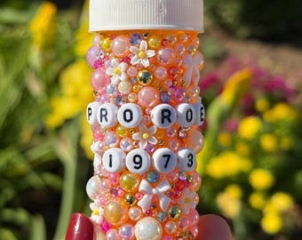 Puede incluir: Un frasco de pastillas naranja decorado con cuentas, flores y lazos rosas, blancos y dorados. El frasco tiene las palabras "PRO RO" y los números "1973" deletreados con cuentas de letras blancas. La tapa del frasco es blanca.
