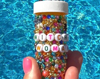Puede incluir: Una botella de pastillas decorada con las palabras "WITCH WOMAN" deletreadas en cuentas de letras. La botella está cubierta de cuentas y gemas de colores. El fondo es una piscina azul brillante.