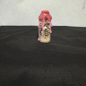 May include: A small glass bottle filled with layers of sand, herbs, and glitter. The bottle is sealed with a pink wax top that has dripped down the sides. The background is a neutral gray and black.
