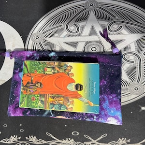 Könnte beinhalten: Ein Tarotkartenspiel mit einer farbenfrohen Illustration liegt auf einem Kordelzugbeutel im Galaxien-Design. Die Tasche ist lila und blau. Ein weißer Halbmond und ein dekoratives kreisförmiges Design befinden sich im Hintergrund.