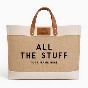 May include: A large tote bag with a tan burlap-like center and off-white top and bottom. The bag has brown leather handles and a leather patch that says "Life's Good". The text "ALL THE STUFF YOUR NAME HERE" is printed on the front.