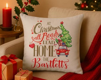 Könnte beinhalten: Ein quadratisches, beiges Weihnachtskissen mit dem Text "At Christmas all Roads Lead Home the Bartlett's". Das Kissen zeigt ein rotes Auto, das einen Weihnachtsbaum und Geschenke transportiert. Ein rotes Herz und Sterne sind ebenfalls auf dem Kissen.