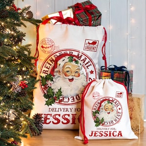 Puede incluir: Dos sacos de Papá Noel de lona blanca con cordones rojos, con un gráfico vintage de Papá Noel y el texto "REINDEER MAIL". El saco más grande está lleno de regalos envueltos. El texto "DELIVERY FOR: JESSICA" está impreso en los sacos.