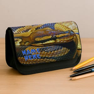 May include: A black pencil case with a detailed image of a snake in brown, yellow, and blue. The text "NAME HERE" is printed in blue on the front. The case is rectangular with a black zip.
