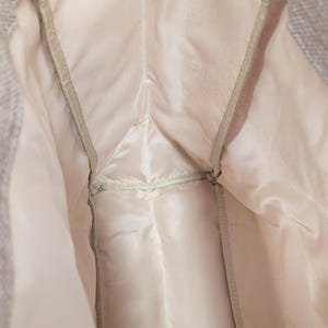 May include: Close-up of the interior of a light-colored garment, possibly a jacket or coat. The lining is a pale pink satin-like material, with seams and a zipper visible. The outer fabric appears to be a textured, light gray material.