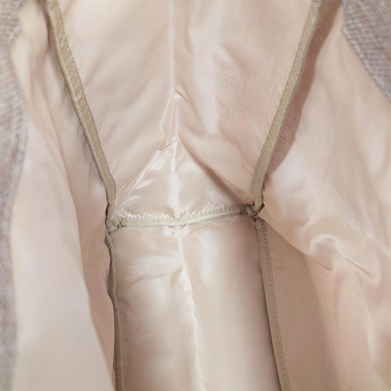 May include: Close-up of the interior of a light-colored garment, possibly a jacket or coat. The lining is a pale pink satin-like material, with seams and a zipper visible. The outer fabric appears to be a textured, light gray material.