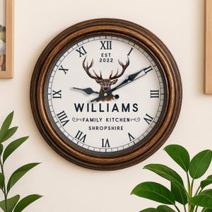 May include: A circular wall clock with a brown and gold frame. The white clock face features black Roman numerals and hands. A deer graphic is above the text "WILLIAMS FAMILY KITCHEN SHROPSHIRE". The year "EST 2022" is above the deer.
