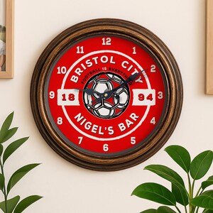 May include: Circular wall clock with a red face, displaying "Bristol City" and a football design. The clock has a dark brown, distressed frame and white numerals. The text "Nigel's Bar" is also visible. The clock's diameter is approximately 30 cm.