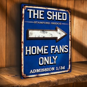 May include: A rectangular blue metal sign with white text that reads "THE SHED - STAMFORD BRIDGE" and "HOME FANS ONLY". An arrow points right. The sign has a distressed, rusty border and is displayed on a wooden shelf.