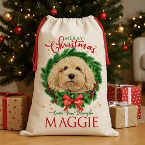 Könnte beinhalten: Ein cremefarbener Weihnachtsgeschenksack mit rotem Kordelzug. Er zeigt ein Hundeporträt in einem grünen Kranz mit roter Schleife. Der Text lautet "Merry Christmas" und "Santa Paws Delivery for MAGGIE".