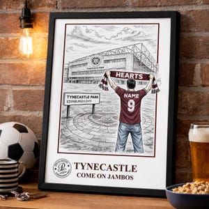 May include: Framed print of Tynecastle Park stadium, featuring a fan with a Hearts scarf. The print includes the text "TYNECASTLE PARK EDINBURGH EH11" and "COME ON JAMBOS". A football and a striped mug are visible.