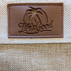 May include: Close-up of a brown leather patch with the words "Life's Good" in cursive script. The patch features a palm tree design and is sewn onto a beige woven fabric.