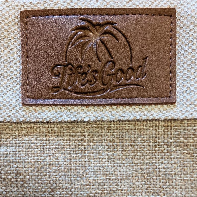 May include: Close-up of a brown leather patch with the words "Life's Good" in cursive script. The patch features a palm tree design and is sewn onto a beige woven fabric.