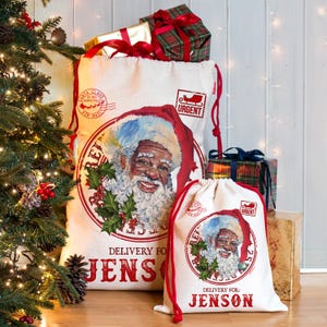 May include: Two white canvas Santa sacks with red drawstring closures. The sacks feature a smiling Santa Claus graphic within a circular frame, with the text "Delivery For: Jenson" and "Urgent" stamps. Presents are visible.