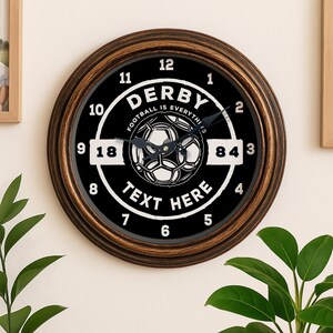 May include: Circular wall clock with a black face and a brown frame. The clock has a soccer ball graphic and the words "DERBY" and "FOOTBALL IS EVERYTHING". The numbers are white, and the hands are black.