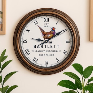 May include: A circular wall clock with a brown and gold frame. The white clock face has black Roman numerals and hands. It features a pheasant illustration and the text "BARTLETT FAMILY KITCHEN SHROPSHIRE EST 2022".