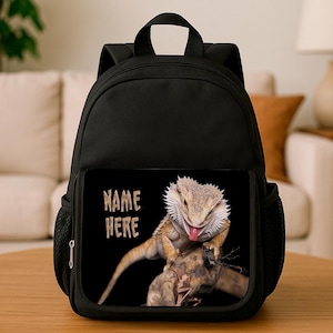 May include: Black backpack with a front pocket displaying a bearded dragon image. The image includes the text "NAME HERE" in white. The lizard is tan, with a pink tongue, and is perched on a piece of wood.