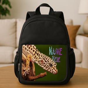May include: Black backpack with a gecko design on the front. The gecko is brown and tan with black spots, on a branch against a green background. The text "NAME HERE" is displayed on the front pocket in a colorful font.