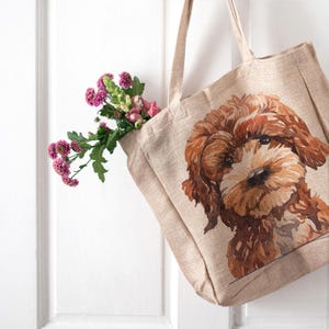 Peut inclure: Sac fourre-tout beige avec une illustration à l'aquarelle d'un chien brun et beige. Le sac contient des fleurs roses et vertes. Le sac fourre-tout a deux poignées et est fait d'un tissu texturé.