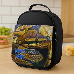 May include: Black lunch bag with a detailed image of a snake's head and body, showcasing brown, yellow, and blue scales. The bag includes a handle and a front pocket with the text "NAME HERE" in blue. The bag is on a wooden table.