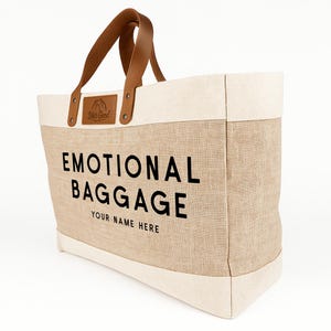 May include: A beige tote bag with the words "EMOTIONAL BAGGAGE" printed in black. The bag has brown leather handles and a cream-colored top and bottom trim. The text "YOUR NAME HERE" is printed below the main text.