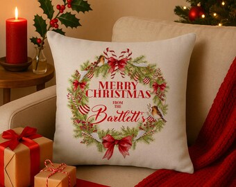 Könnte beinhalten: Ein quadratisches, cremefarbenes Weihnachtskissen mit einem Kranzdesign. Der Kranz enthält rote Schleifen, Zuckerstangenstreifen und Vögel. Der Text "MERRY CHRISTMAS from the Bartlett's" ist rot gedruckt. Das Kissen liegt auf einem beigen Sofa.