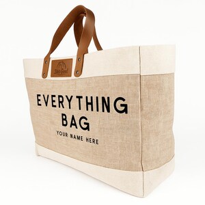 May include: A beige canvas tote bag with the words "EVERYTHING BAG" in large black letters. The bag has cream-colored trim and brown leather handles. The text "YOUR NAME HERE" is below the main text. A small leather patch reads "Life's Good".