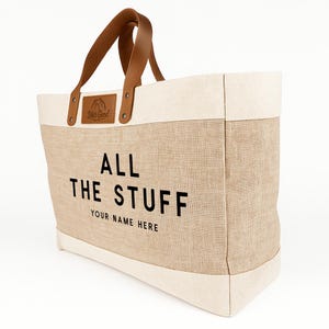 May include: A large tote bag with a natural beige canvas body and off-white trim. The bag features brown leather handles and a leather patch with the text "Life's Good". The words "ALL THE STUFF" and "YOUR NAME HERE" are printed in black.