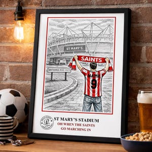May include: Framed black and white print of St. Mary's Stadium with a person in a Saints jersey holding a scarf. The print includes the text "ST MARY'S STADIUM OH WHEN THE SAINTS GO MARCHING IN."