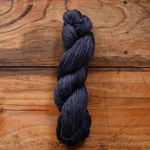 May include: A skein of dark gray yarn, tightly twisted, against a wooden background. The yarn appears soft and has a slight sheen, suggesting a blend of fibers. The color is a deep, neutral gray, suitable for various knitting or crochet projects.