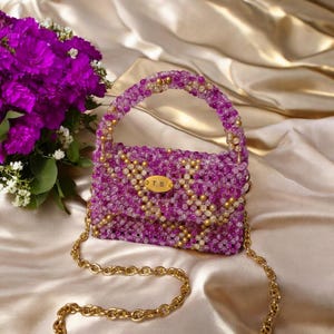 Könnte beinhalten: Eine perlenbesetzte Handtasche in Lila und Gold, mit einem goldenen Kettenriemen und Griff. Die Tasche ist mit kleinen Perlen verziert, mit einer goldenen Plakette mit der Aufschrift "T.S.". Die Tasche steht vor einem Satinhintergrund, mit einem Strauß lila Blumen.