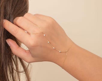 May include: Delicate gold hand chain bracelet featuring small, round, white beads. The bracelet connects to a finger ring and wraps around the wrist. The jewelry is displayed against a neutral background.