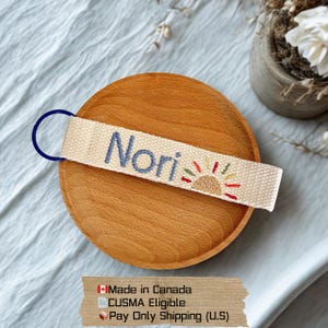 Può includere: Un portachiavi in tessuto beige con il nome "Nori" ricamato in blu e un disegno di sole colorato. Il portachiavi ha un anello di metallo blu e poggia su un piatto di legno.