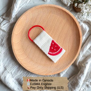Puede incluir: Un llavero blanco con un diseño de rodaja de sandía roja y un lazo rojo. El llavero está sobre un plato de madera. El texto "Made in Canada" y "CUSMA Eligible" son visibles.