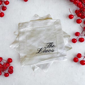 Könnte beinhalten: Ein Stapel cremefarbener Leinen-Servietten mit gesticktem Text "The Liaos" in schwarzer Schrift. Die Servietten haben eine dekorative Naht. Rote Beeren sind auf einer weißen Oberfläche um die Servietten angeordnet.