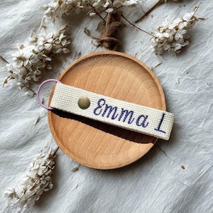 Puede incluir: Llavero de tela color crema con el nombre "Emma 1" bordado en hilo morado. El llavero tiene un broche de color latón y un llavero de color lavanda. El llavero descansa sobre un plato de madera, con flores blancas en el fondo.