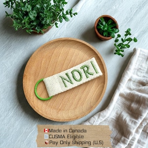 Może przedstawiać: Brelok z białej tkaniny z napisem "NORI" wyhaftowanym zielonymi literami. Brelok ma zielony metalowy pierścień i jest prezentowany na drewnianym talerzu. Tło obejmuje zieleń i białą tkaninę.