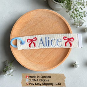 May include: A personalised keychain with the name "Alice" embroidered in blue, with red bow designs on either side. The keychain is made of off-white fabric and has a light blue ring. It sits on a wooden plate.