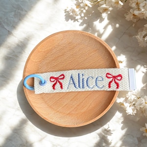 Op de afbeelding: Een crèmekleurige sleutelhanger met de naam "Alice" in blauw geborduurd, met rode strikaccenten. De sleutelhanger heeft een blauwe ring en een zilveren metalen uiteinde. Hij ligt op een ronde houten plaat.