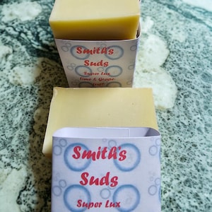May include: Four bars of light yellow soap are stacked in front of a white and blue box. The box has the text "Smith's Suds Super Lux Lime & Grapefruit". The soap is on a green and gray marble surface.