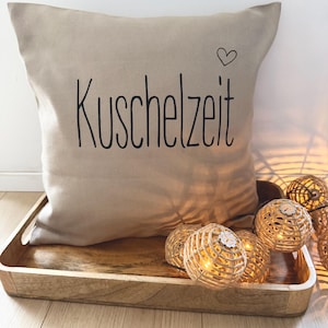 May include: A beige square pillow with the word "Kuschelzeit" in black script and a small heart. The pillow is on a wooden tray with decorative string lights.