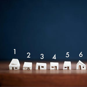 May include: Seven miniature white houses with different designs, numbered 1 through 7, on a wooden surface. A silver coin is to the right of the houses.