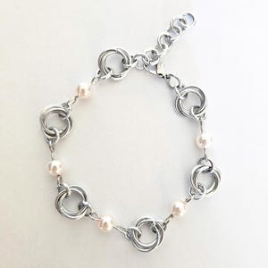 May include: A silver-toned bracelet featuring a series of interlocking circular links. Small, pale pink pearls are interspersed between the links, adding a touch of elegance. The bracelet has an adjustable chain for a comfortable fit.
