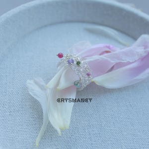 Puede incluir: Un anillo de hilo de plata tejido con pequeñas piedras preciosas facetadas multicolores en tonos rosa, morado, verde y azul. La palabra CRYSMANCY está impresa en negro debajo del anillo, que descansa sobre un pétalo de flor rosa pálido.