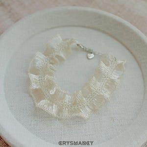 Peut inclure: Un bracelet en dentelle à volants de couleur crème présentant une texture de tissu plissé délicate. L'accessoire comprend une fermeture à chaîne en métal argenté avec une petite breloque en forme de cœur. Le mot CRYSMANCY est imprimé en gris foncé.