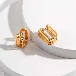 Könnte beinhalten: Ein Paar goldfarbene Creolen. Die Ohrringe haben eine rechteckige Form mit einem geschichteten Design. Die Ohrringe werden auf einer weißen Oberfläche präsentiert.