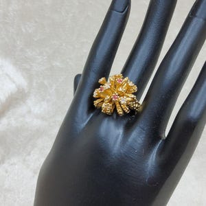May include: A gold ring featuring a textured, multi-layered floral or starburst design. The center of the ring is adorned with small, embedded clear stones and tiny pink gemstones, creating a cluster effect on the metallic gold surface.
